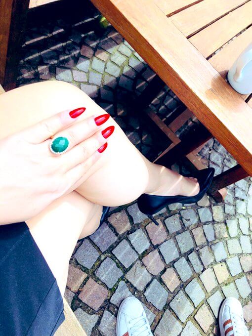 Eine Hand mit langen rot lackierten Fingernägeln und einem Ring mit einem grünen Edelstein liegt auf einem nackten Bein, fertig zur Keuschhaltung.