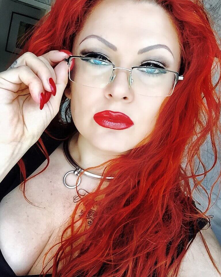 Eine Frau mit leuchtend roten Haaren und Lippen sowie Fingernägeln schaut direkt in die Kamera, damit jemand einen Blackmail-Vertrag unterschreibt, während sie mit der rechten eine randlose Brille auf ihre Nasenspitze schiebt.