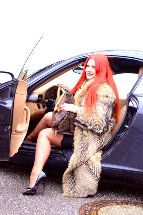Eine Geldherrin mit langen roten Haaren, roten Lippen, Pelzmantel und High Heels steigt mit einer Designertasche aus einem Luxus-Auto.