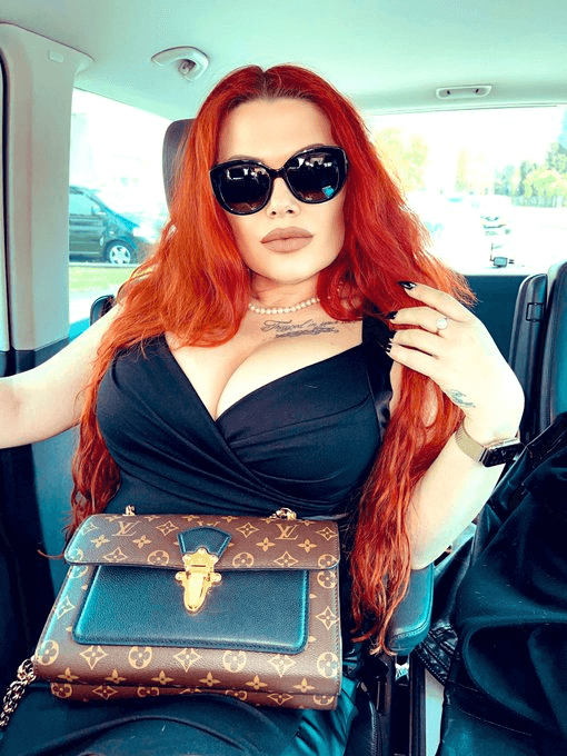 Die Moneydom mit langen roten Haaren sitzt auf der Rückbank ihres Autos mit einem schwarzen, tief ausgeschnittenem Kleid und einer großen schwarzen Sonnenbrille bekleidet und einer Louis Vuitton Tasche auf ihrem Schoß.